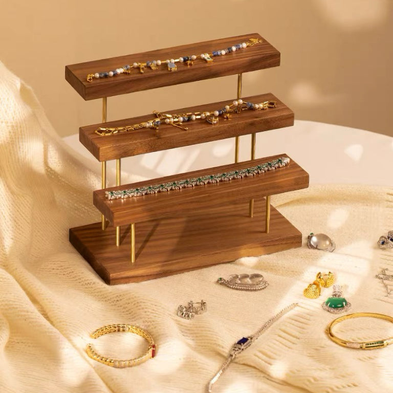 A three-tier wooden jewelry display stand with gold-tone metal legs, styled on a cream fabric surface with various gold, silver, and multicolor jewelry pieces arranged around it.