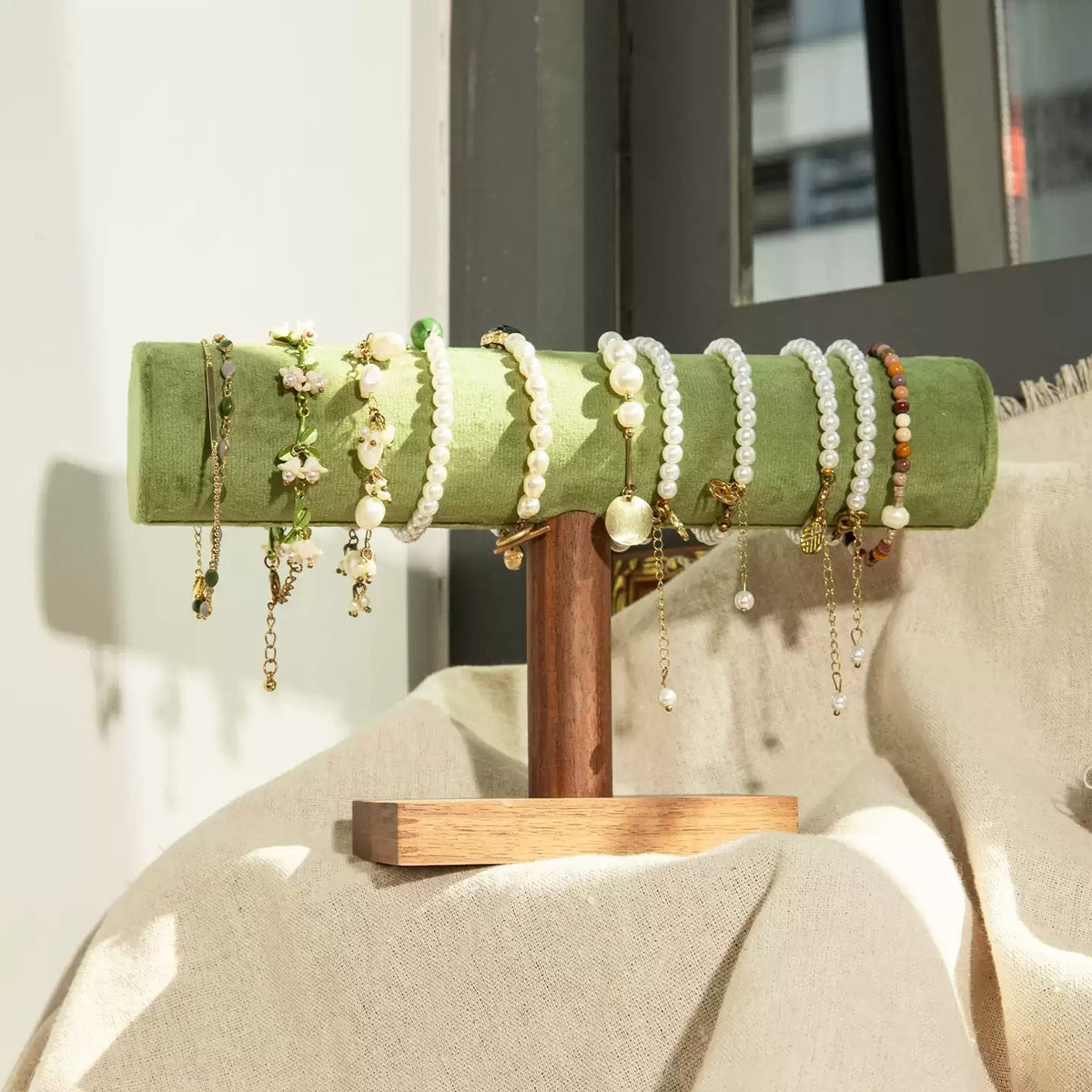 A set of beaded bracelets and delicate jewelry displayed on a green velvet jewelry stand with a wooden base, set against a draped beige fabric background.