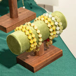 A set of beaded bracelets displayed on a green velvet cylindrical jewelry stand mounted on a wooden base. The bracelets feature round beads in pale yellow, light green, and white, accented with small gold spacer beads and charms.