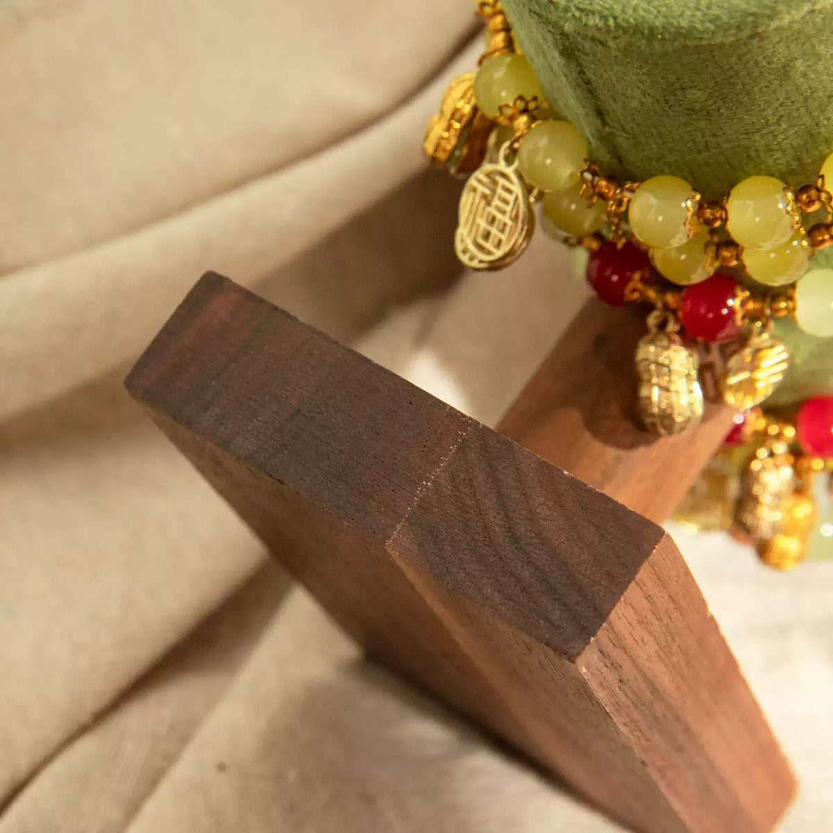 Close-up of a beaded bracelet with gold charms and pale green and red beads displayed on a wooden stand against a beige fabric background.