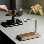 A wooden and metal coffee accessory set on a gray tabletop, including a wooden scoop being used to pour coffee beans into a scale, a metal ring holder, and a wooden stand in the foreground.