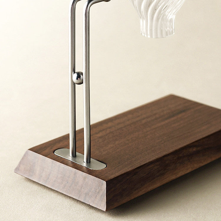 A close-up of a modern table lamp base featuring a dark brown wood block with a triangular front, a polished silver metal stand with a small ball accent, and a clear ribbed glass shade at the top, set against a warm beige background.