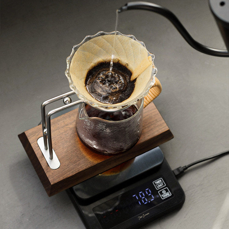 A pour-over coffee setup with dark coffee dripping into a glass carafe placed on a digital scale, with a metal handle and wooden base, against a gray background.