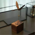 A wooden bird-shaped wind spinner ornament mounted on a square wooden base, photographed on a tabletop with a softly blurred indoor background.