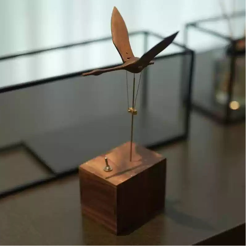 A wooden bird-shaped wind spinner ornament mounted on a square wooden base, photographed on a tabletop with a softly blurred indoor background.