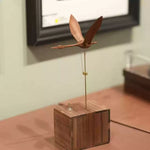 A wooden bird wind spinner toy with a natural wood grain finish, mounted on a vertical metal rod over a square wooden base, photographed indoors against a softly blurred background.