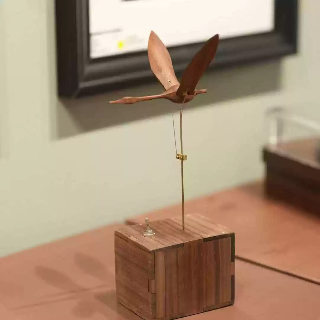 A wooden bird wind spinner toy with a natural wood grain finish, mounted on a vertical metal rod over a square wooden base, photographed indoors against a softly blurred background.