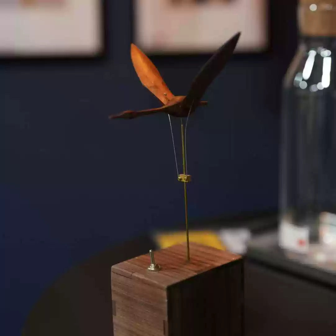 A wooden bird wind spinner mounted on a square wooden base, photographed indoors against a dark blue wall with a blurred glass bottle in the background.