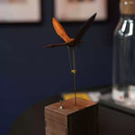 A wooden bird wind spinner mounted on a square wooden base, photographed indoors against a dark blue wall with a blurred glass bottle in the background.