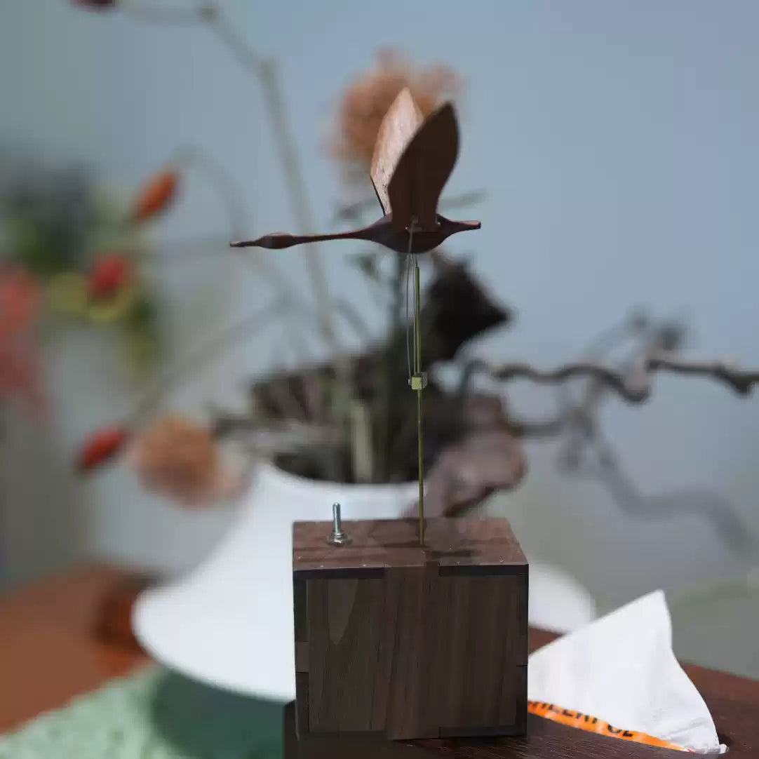 A wooden bird wind spinner on a dark wood base, photographed on a tabletop with a blurred indoor background and dried flowers.