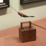 A wooden mechanical toy or sculpture with a rectangular base, small handle, and thin wire arms supporting a curved wooden top, displayed on a tabletop in an indoor setting.