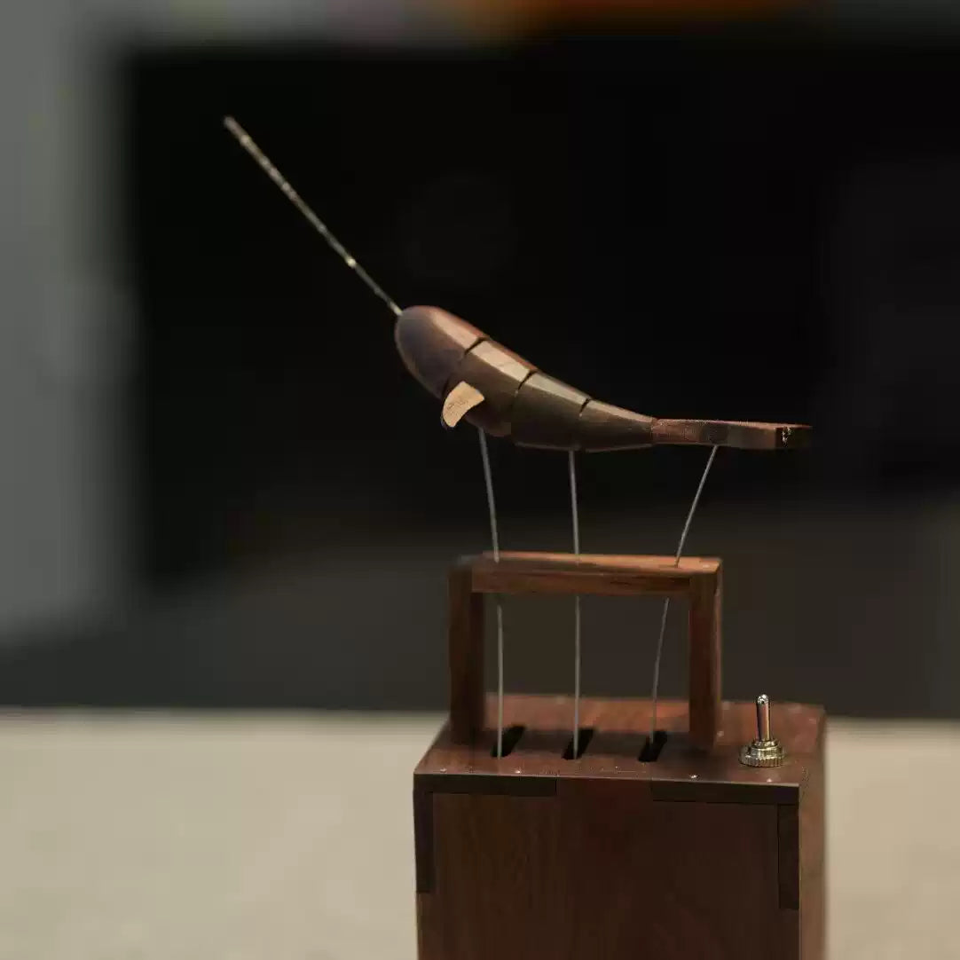 A wooden and metal mechanical bird sculpture mounted on a wooden base, photographed against a dark blurred indoor background.