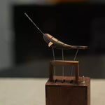 A wooden and metal mechanical bird sculpture mounted on a wooden base, photographed against a dark blurred indoor background.