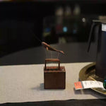 A small wooden fish-shaped ornament or figurine mounted on a square wooden base, displayed on a light fabric surface with a dark blurred background and part of a black pitcher visible at the right edge.