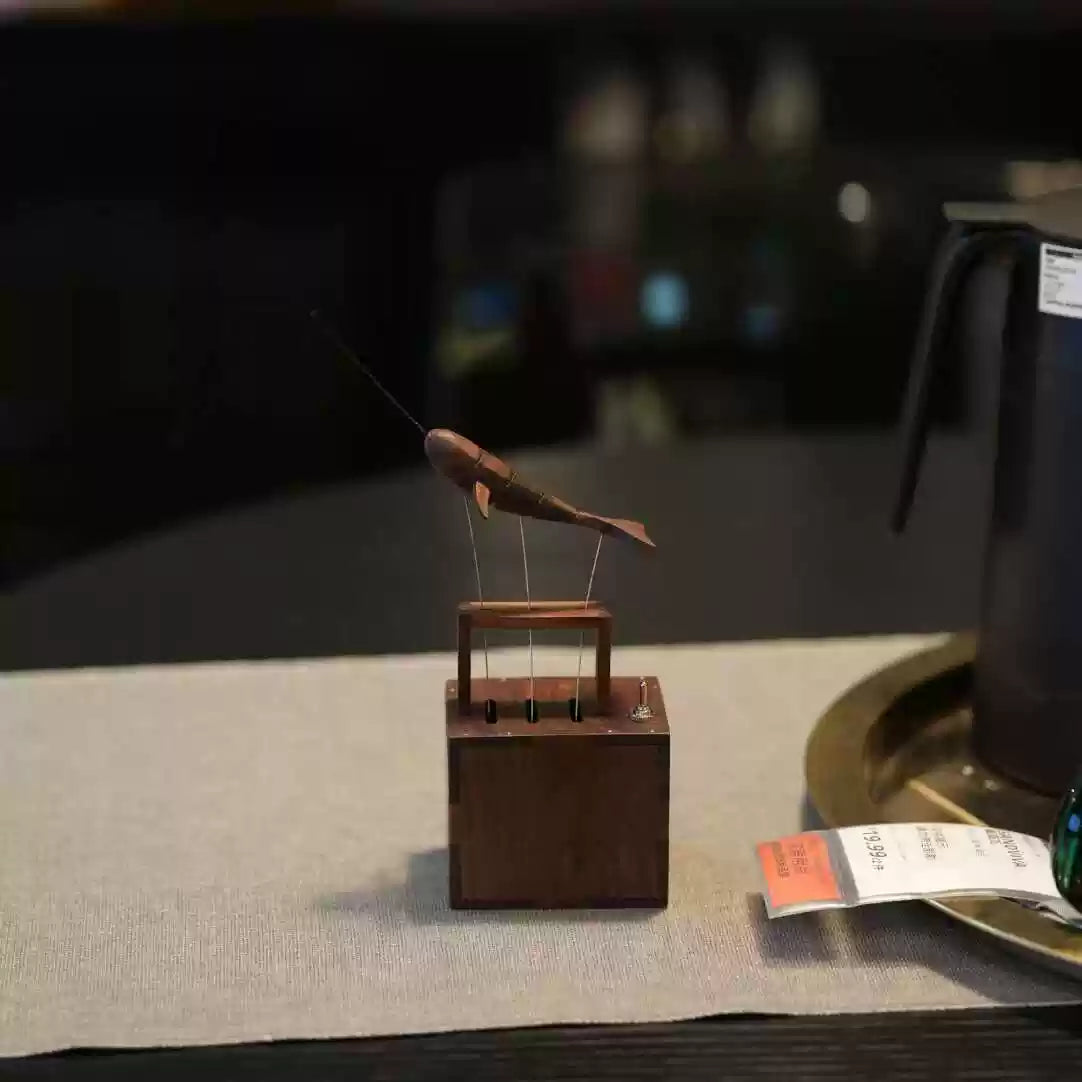 A small wooden fish-shaped ornament or figurine mounted on a square wooden base, displayed on a light fabric surface with a dark blurred background and part of a black pitcher visible at the right edge.