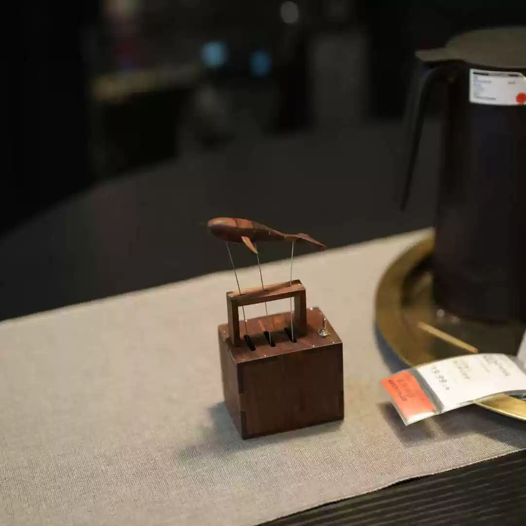 A small wooden display box holding a fish-shaped object on thin metal wires, placed on a light fabric surface in a dim indoor setting. A dark vessel and a small orange-and-white tag are partially visible in the background.