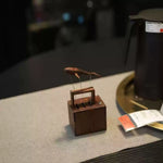 A small wooden display box holding a fish-shaped object on thin metal wires, placed on a light fabric surface in a dim indoor setting. A dark vessel and a small orange-and-white tag are partially visible in the background.
