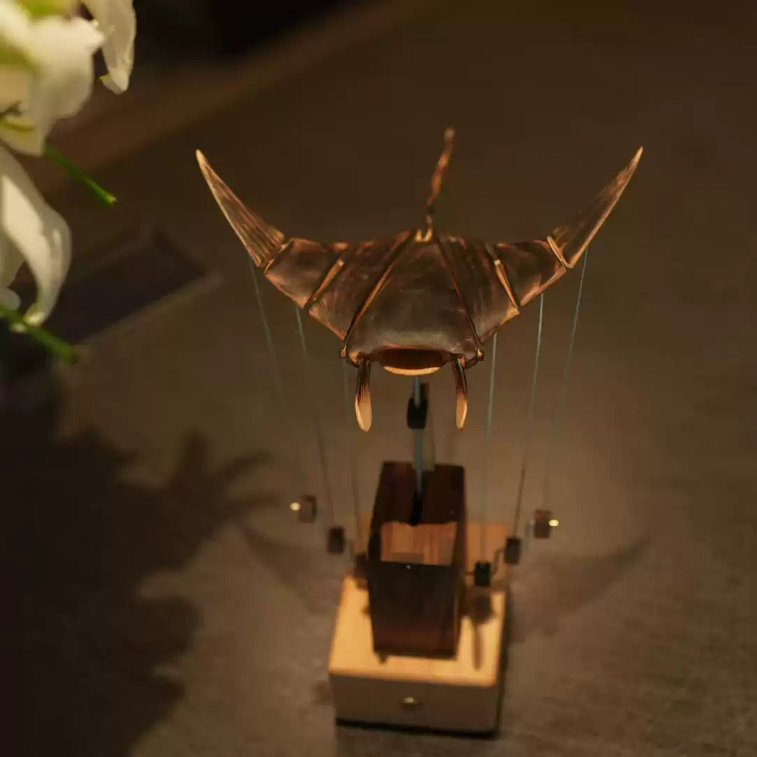 A bronze insect sculpture mounted on a small wooden base, photographed in a dim indoor setting with warm lighting and soft shadows.