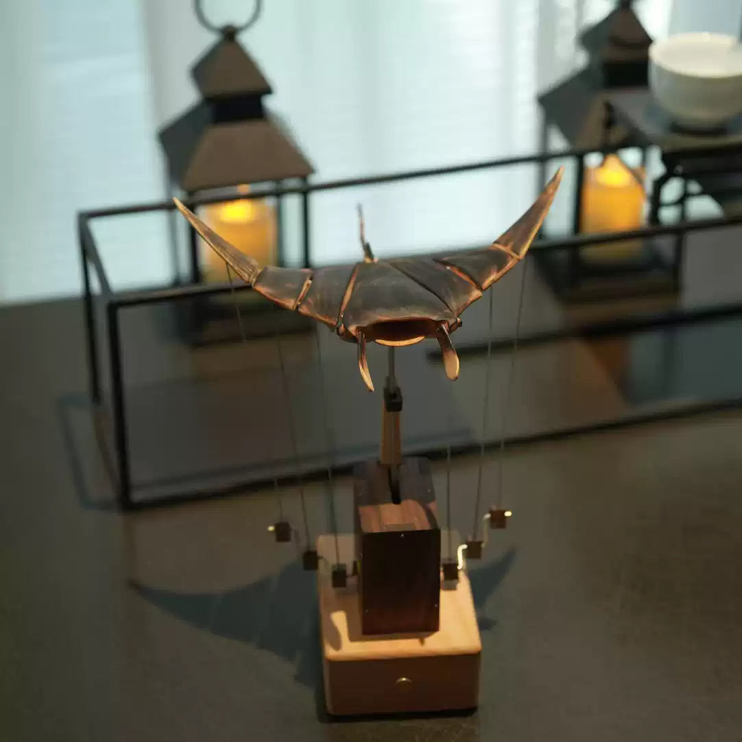 A marionette-style puppet toy with a metallic winged figure suspended by strings on a wooden base, photographed on a tabletop with blurred lanterns in the background.