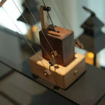 A close-up of a wooden string instrument component, likely a harp or zither, featuring a dark brown wooden block, a light natural wood base, and thin metal strings, set against a softly blurred indoor background.