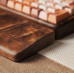 A close-up of a wooden keyboard wrist rest with a rich dark brown finish, resting on a textured beige fabric next to a mechanical keyboard with warm-toned keycaps.