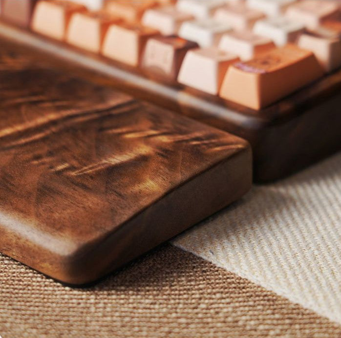 A close-up of a wooden keyboard wrist rest with a rich dark brown finish, resting on a textured beige fabric next to a mechanical keyboard with warm-toned keycaps.