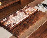 A wooden mechanical keyboard with a matching wood wrist rest, featuring warm brown, beige, and cream keycaps with small character illustrations, set on a woven desk mat.