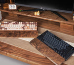 A wooden desk organizer setup featuring a black mechanical keyboard on a dark wood tray, a brown and beige patterned keyboard on a matching tray, and a black keyboard on a wood-grain wrist rest, all arranged under a computer monitor.