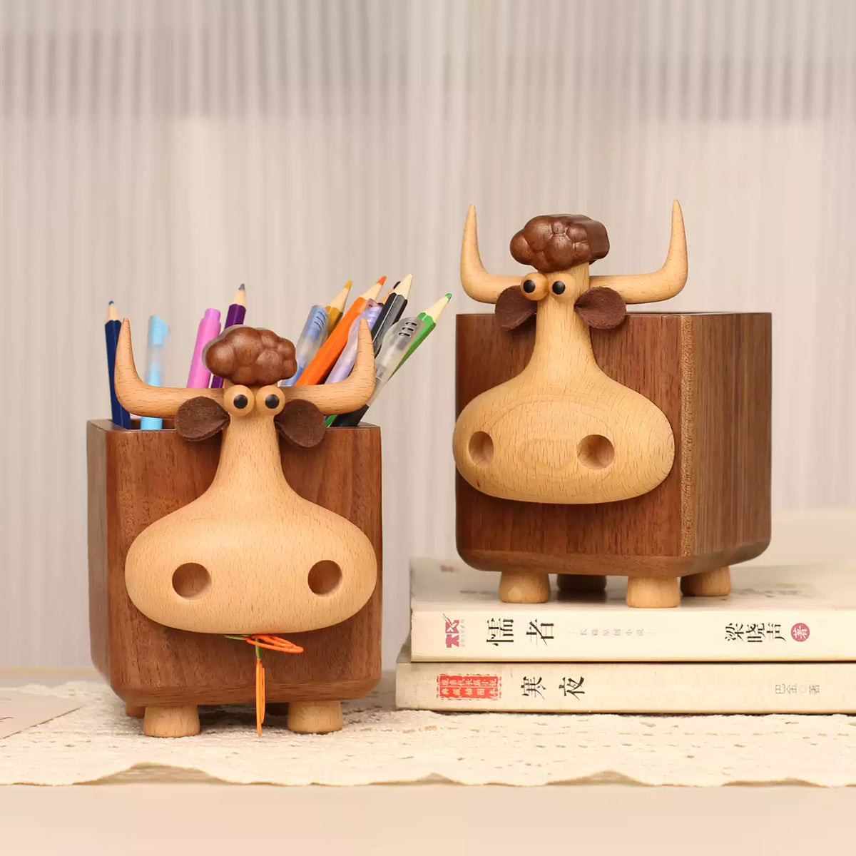 Two wooden cow-shaped pencil holders on a tabletop, one holding colored pencils and the other empty, styled with books against a soft neutral background.