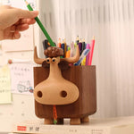 A wooden cow-shaped pencil holder with a carved wood nose and horns, filled with colored pencils and held by a hand placing a green pencil, set on a desk with a book and white curtain background.