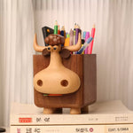 A wooden cow-shaped pencil holder with a light natural wood face and darker brown body, filled with colorful pencils, sitting on top of books against a soft cream curtain background.