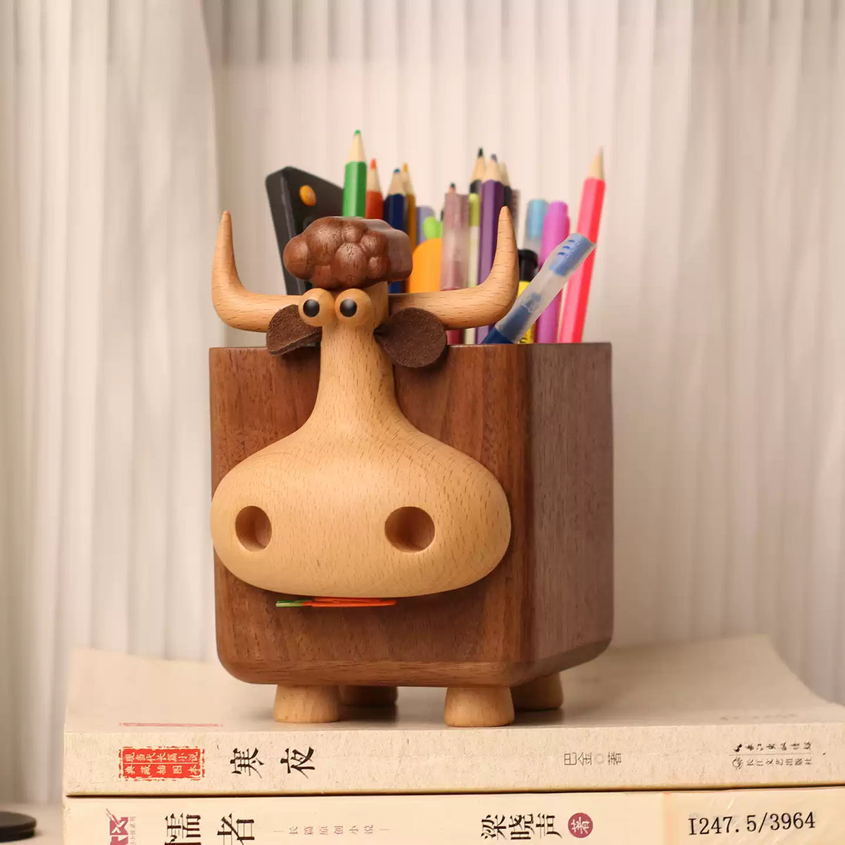 A wooden cow-shaped pencil holder with a light natural wood face and darker brown body, filled with colorful pencils, sitting on top of books against a soft cream curtain background.