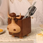 A wooden cow-shaped utensil holder with a carved wooden nose and horns, holding metal chopsticks and a fork, set on a lace doily with a blurred kitchen background.