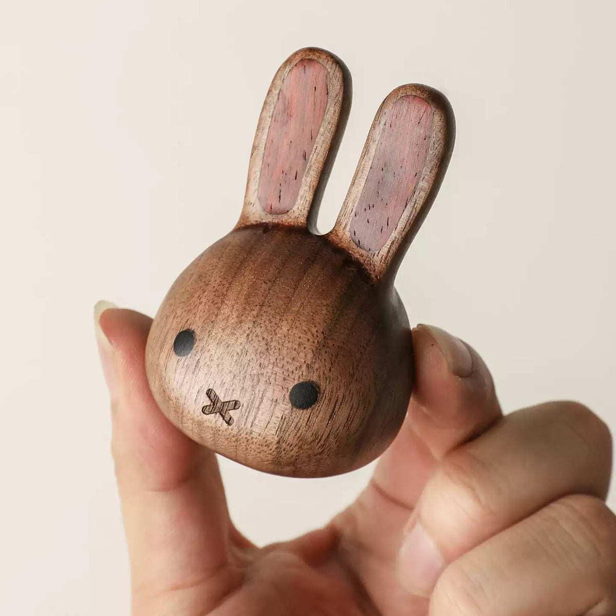 A hand holding a small wooden rabbit-shaped object with long ears, dark round eyes, and a simple stitched mouth, shown against a plain light background.