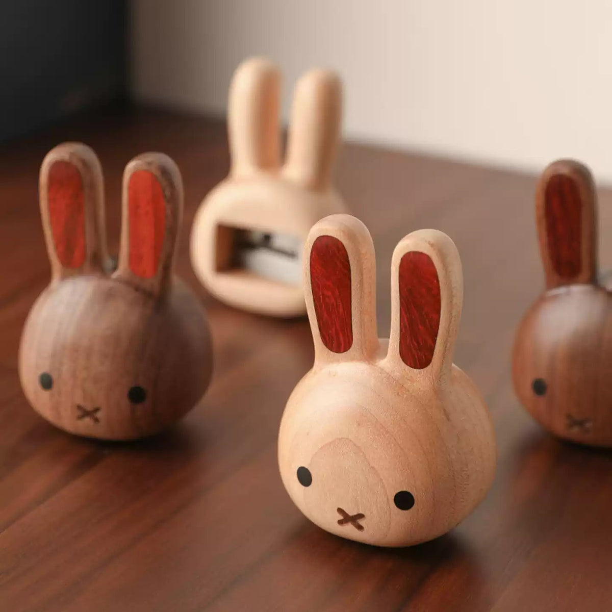 Four small wooden rabbit-shaped objects with long ears and simple facial details are arranged on a wooden tabletop. The center rabbit is in focus, while the others are softly blurred in the background.