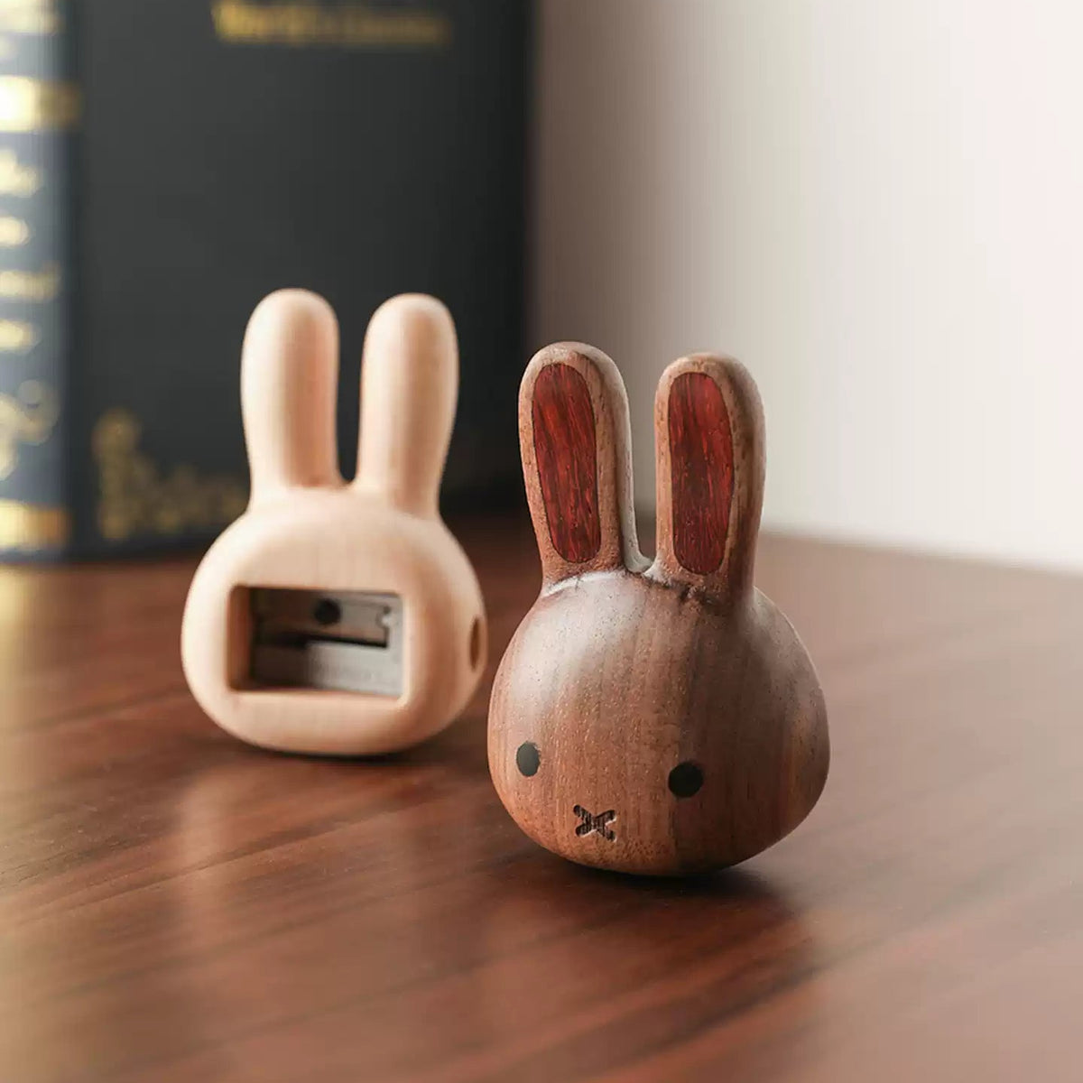 Two wooden rabbit-shaped pencil cases on a wooden table, one in light natural wood and one in medium brown wood, with a blurred book in the background.
