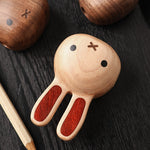 A wooden rabbit-shaped object with a light natural wood head and ears inlaid with reddish wood, photographed on a dark wood surface. Part of a matching pencil-like tool is visible at the lower left, and other similar wooden items appear partially cropped at the top.